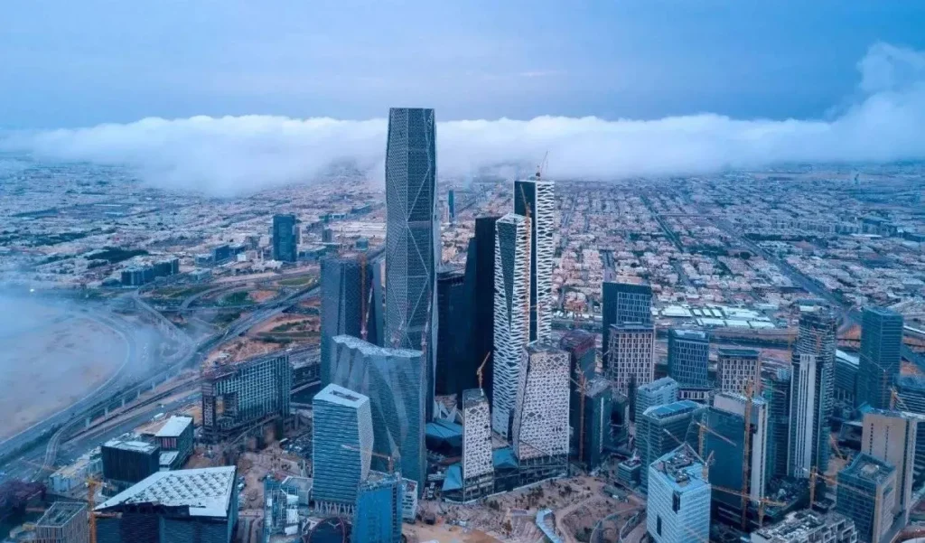A wide-angle landscape shot of the Riyadh skyline at sunset, featuring the illuminated Kingdom Centre tower and modern skyscrapers under a colorful purple and orange sky.