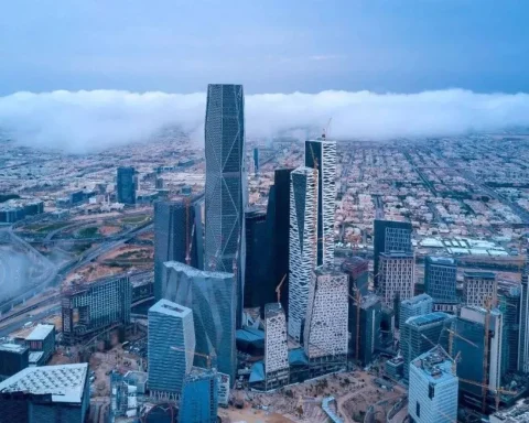 A wide-angle landscape shot of the Riyadh skyline at sunset, featuring the illuminated Kingdom Centre tower and modern skyscrapers under a colorful purple and orange sky.