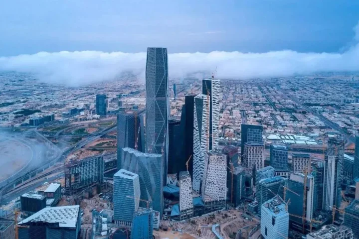 A wide-angle landscape shot of the Riyadh skyline at sunset, featuring the illuminated Kingdom Centre tower and modern skyscrapers under a colorful purple and orange sky.