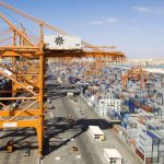 High-angle view of a bustling container terminal at an Omani port, featuring large orange gantry cranes loading a cargo ship alongside rows of stacked shipping containers under a clear blue sky.