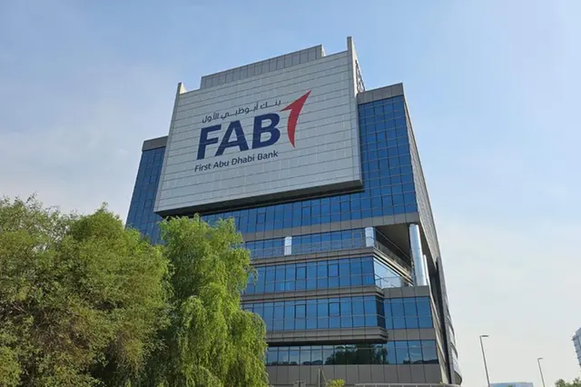 A low-angle shot of the First Abu Dhabi Bank (FAB) headquarters, a modern blue glass-paneled building featuring the large FAB logo against a clear blue sky.